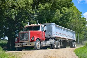 Red and white semi-truck with a large gray trailer driving on a gravel road with trees and blue sky in the background.
